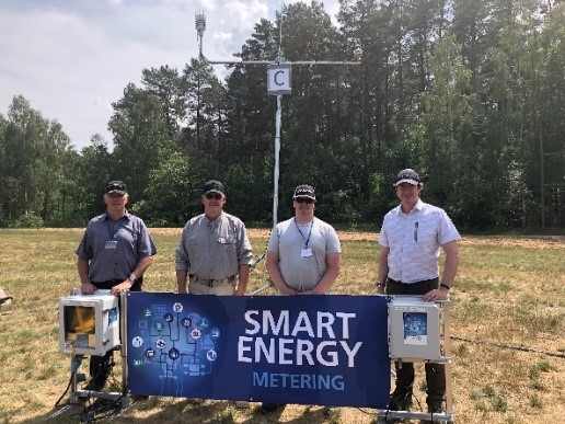 Quatre collègues masculins sont debout dans un champ derrière une banderole sur laquelle on peut lire « Smart energy metering ». On y voit aussi les outils développés au cours du projet.