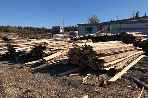 Cour à bois renfermant de longs morceaux de déchets de bois empilés devant un bâtiment industriel.