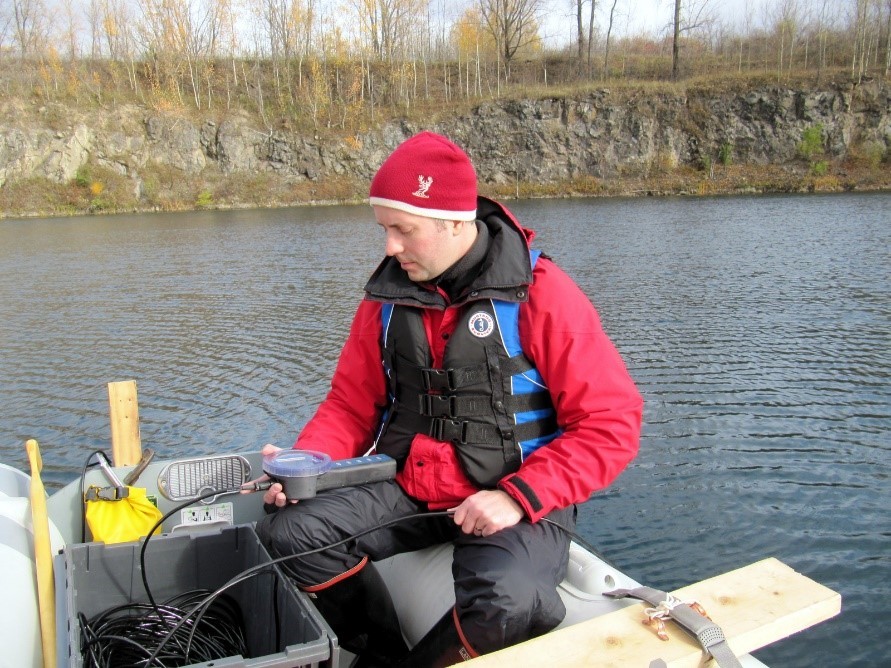 Michael Parsons est assis dans un bateau au milieu d’un plan d’eau, un appareil de mesure à la main servant à déterminer la qualité de l’eau d’une ancienne mine.